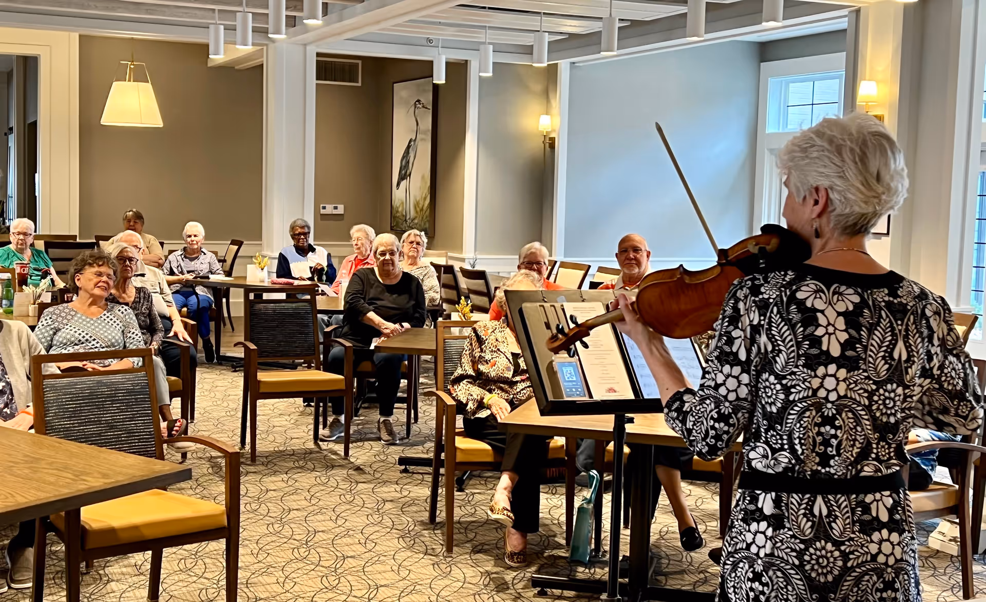 A group of elderly people seated at tables in a well-lit room attentively watching a woman playing the violin. The room has beige walls, patterned carpet, and framed artwork on the walls.