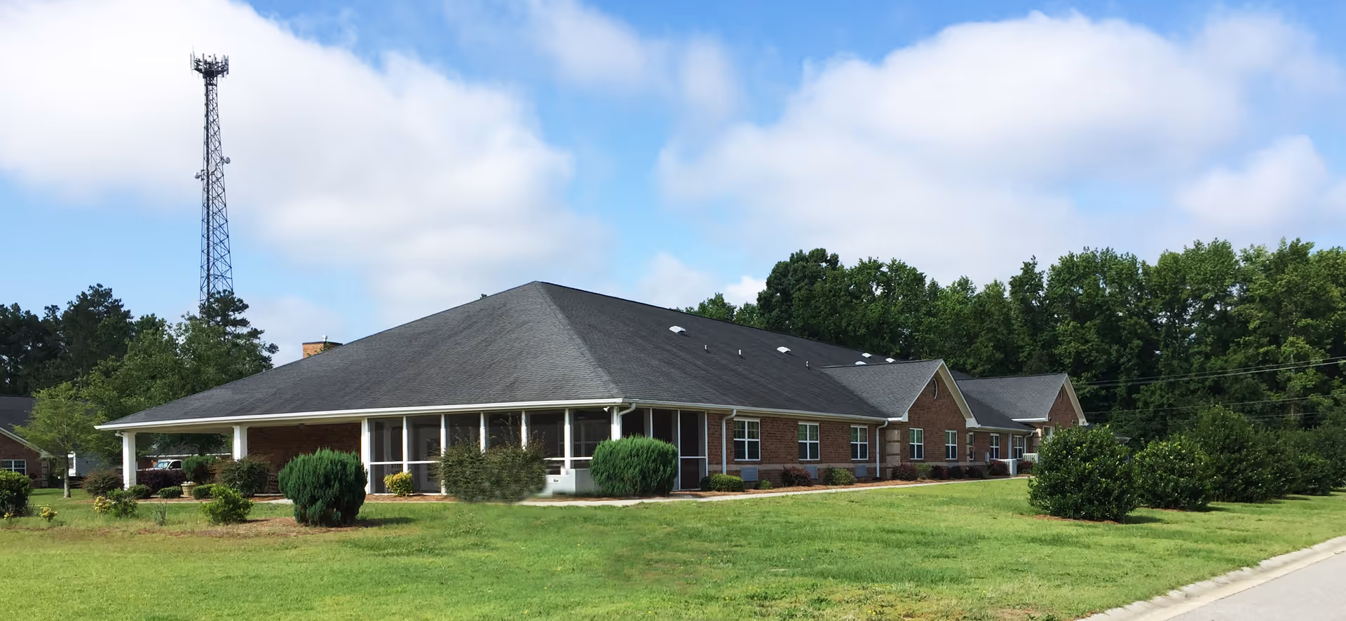 Single-story brick building with a dark gray roof surrounded by green grass and bushes under a partly cloudy blue sky. A tall communication tower is visible in the background.