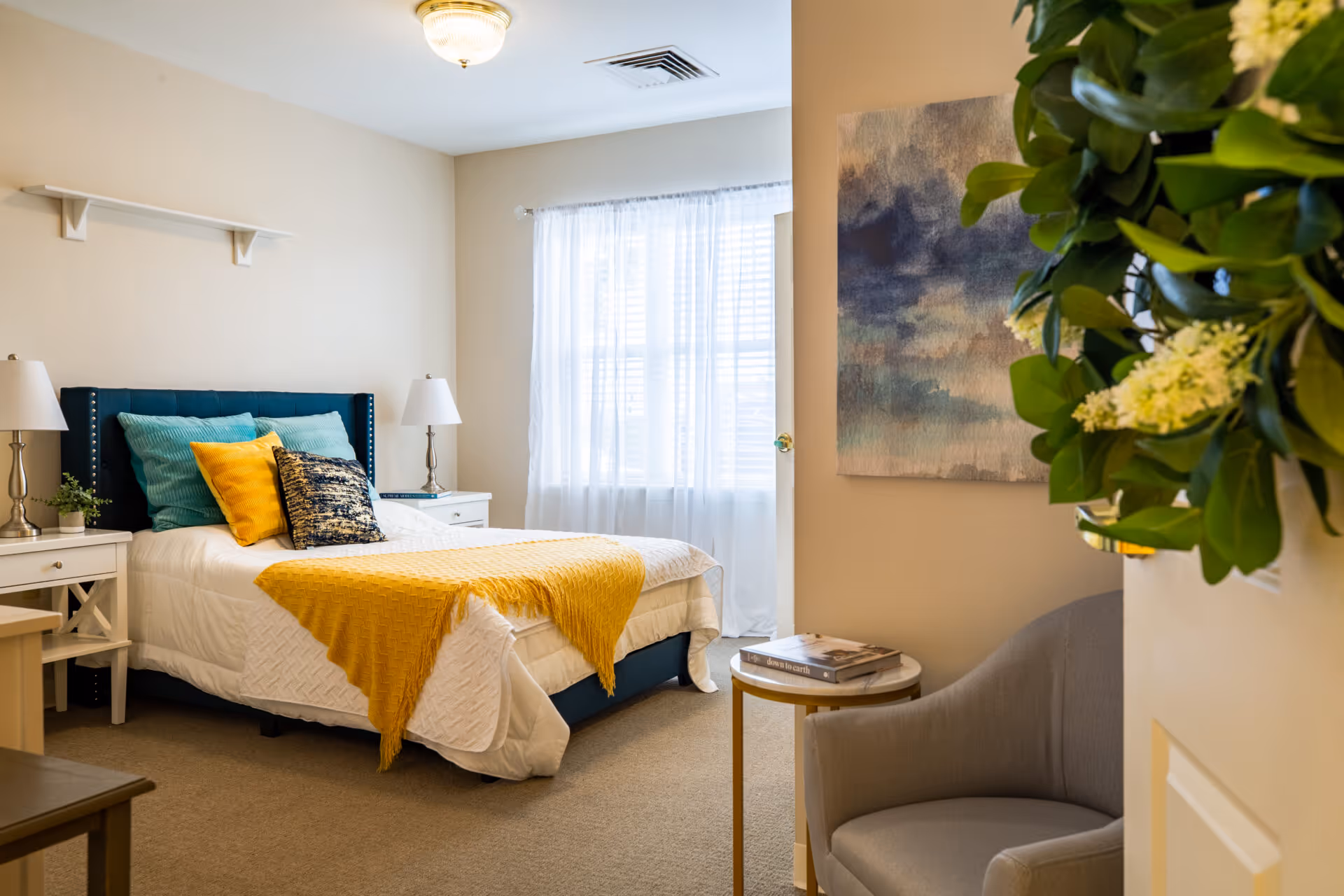 A cozy bedroom with a neatly made bed featuring a white quilt, teal and yellow pillows, and a yellow throw blanket. There are two white nightstands with matching lamps on either side of the bed. A window with sheer white curtains allows natural light to fill the room. A gray armchair and a small round side table with books are positioned near the entrance. A large abstract painting hangs on the wall, and a green leafy plant is partially visible in the foreground.