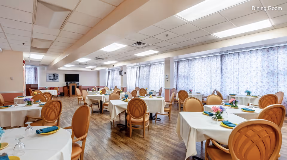 A spacious dining room with multiple round tables covered with white tablecloths, each set with plates, napkins, glasses, and small flower arrangements. The room has wooden flooring, large windows with patterned curtains, and a television mounted on the far wall.
