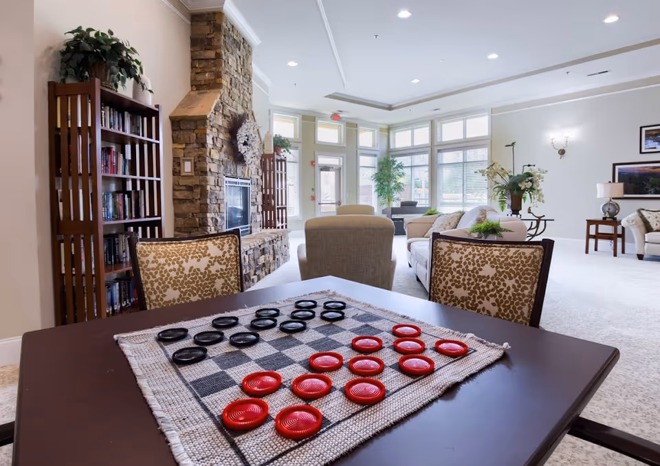 A bright and spacious living room area with large windows letting in natural light. In the foreground, there is a table with a checkers game set up with red and black pieces. The room features comfortable upholstered chairs and sofas, a stone fireplace with a decorative wreath, bookshelves filled with books, and various plants and framed artwork on the walls.