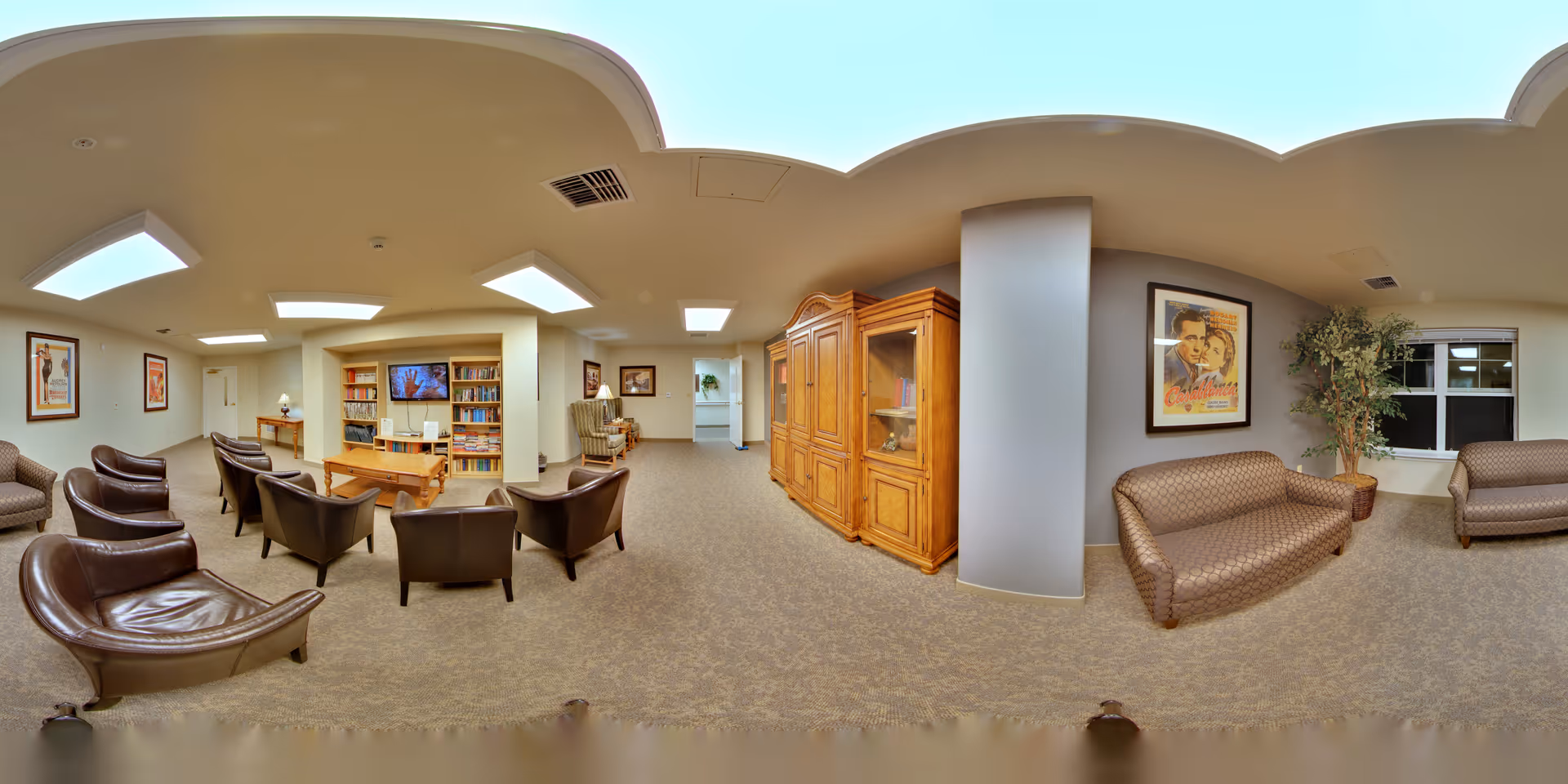 A spacious senior living facility common area with multiple seating options including leather chairs and patterned sofas. There is a wooden cabinet, a bookshelf filled with books, a flat-screen TV mounted on the wall, framed artwork, and a potted plant near a window. The room is well-lit with ceiling lights and has a carpeted floor.