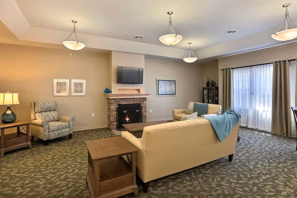 Cozy senior living common room with sofas and armchairs arranged around a fireplace and wall-mounted TV, pendant lights, and large curtained windows.