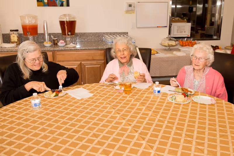 Three elderly women sitting around a table with a checkered tablecloth, eating food and drinking water in a room with a countertop and beverage dispensers in the background.