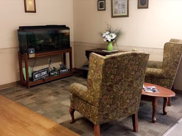 Small sitting area with two patterned armchairs, a round coffee table, a fish tank on a wooden stand, and a vase of flowers on a side table.
