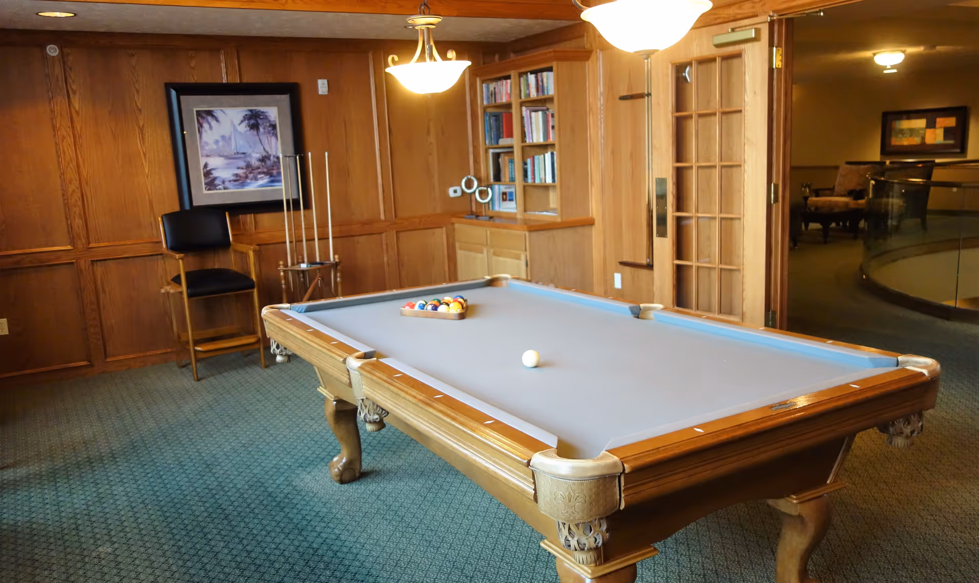 A cozy game room with a wooden pool table set up with billiard balls and a cue ball. The room has wood-paneled walls, a framed painting, a tall chair, a rack holding pool cues, a built-in bookshelf filled with books, and a glass-paned door leading to another area with additional seating.