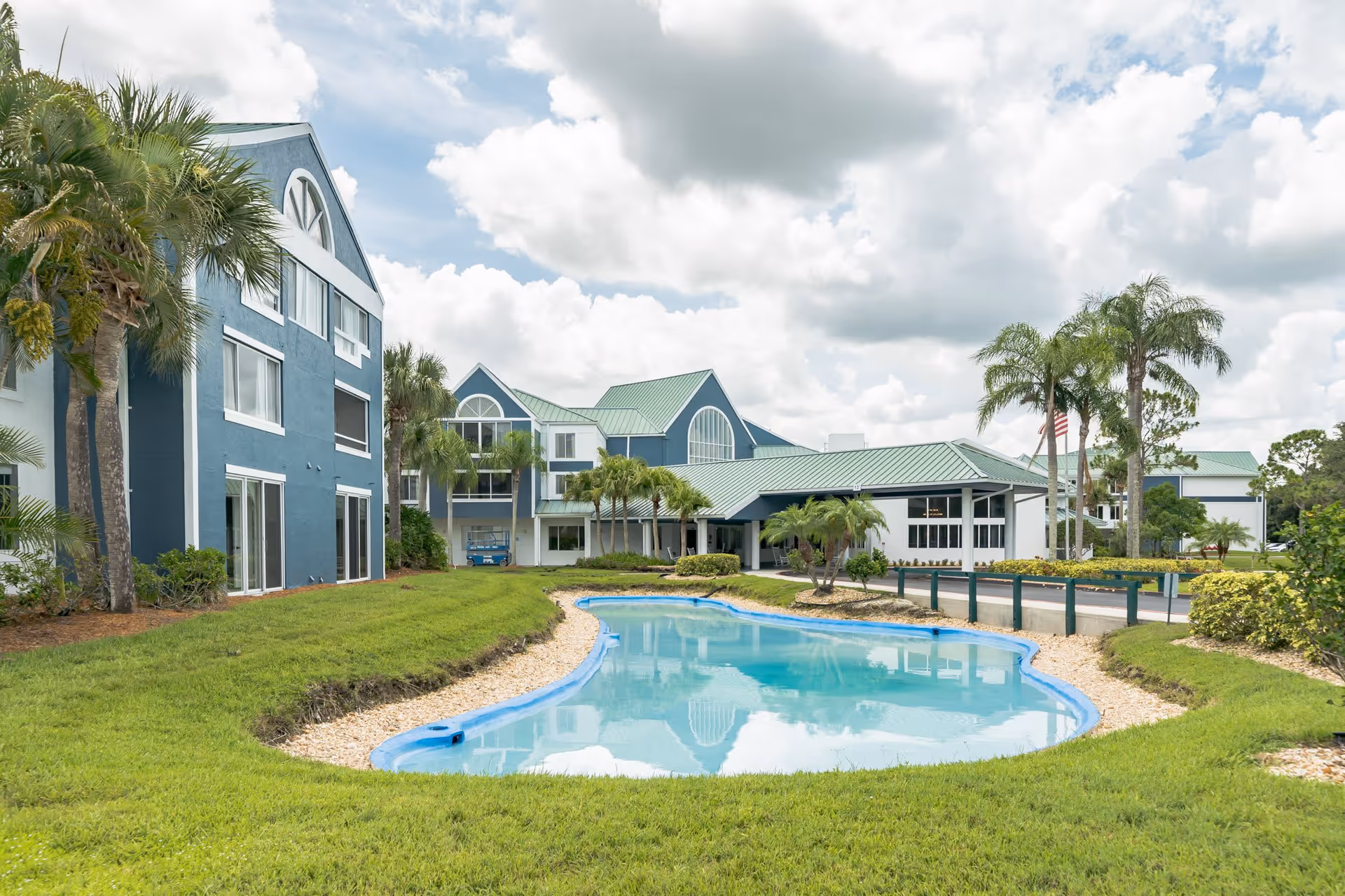 Exterior view of Discovery Village Vero Beach facility featuring blue and white buildings with green roofs, palm trees, a small pond, and a partly cloudy sky.