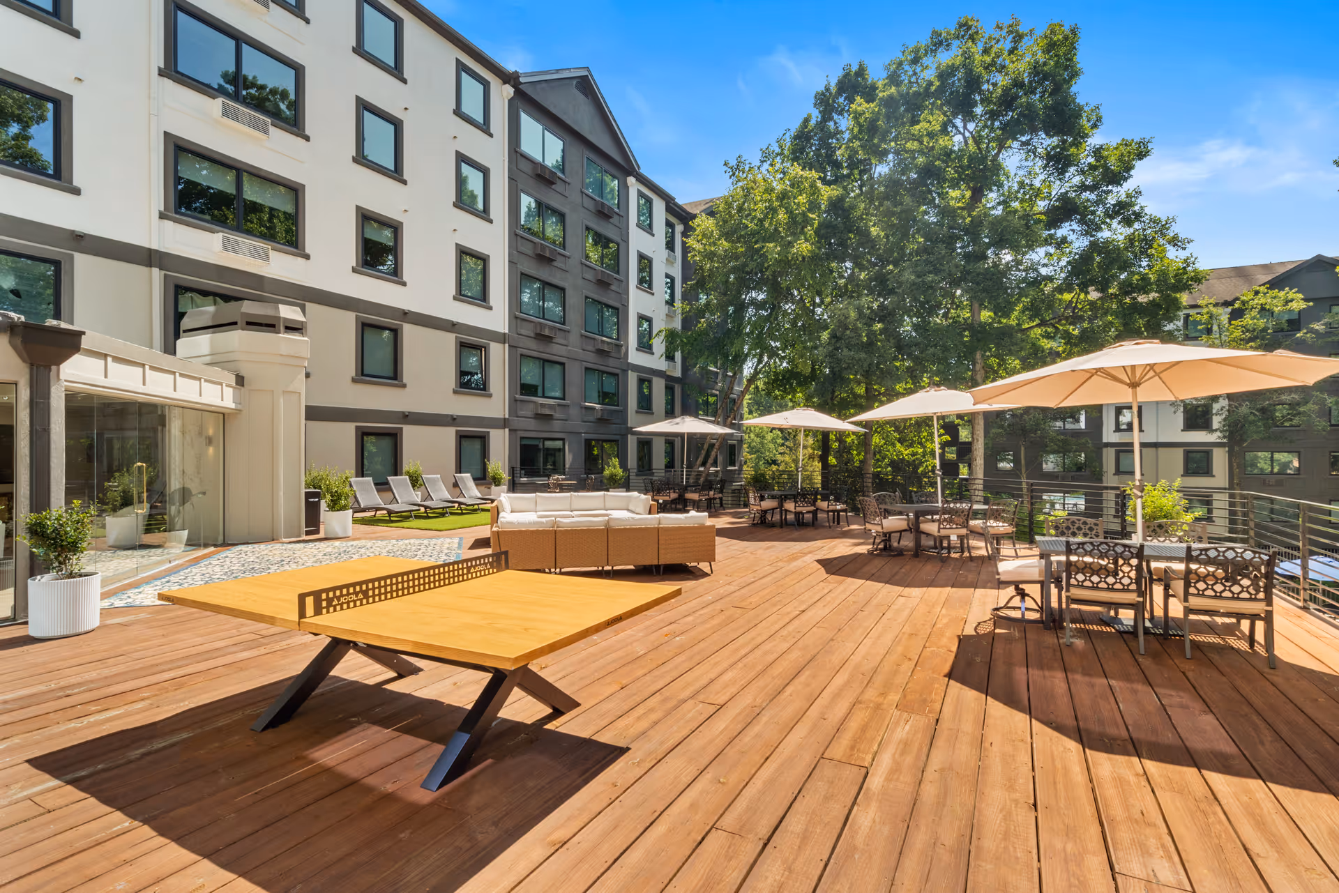Outdoor wooden deck area at Clairmont Crest - 55+ Active Adult Community with tables, chairs, umbrellas, a ping pong table, lounge chairs, and trees in the background under a clear blue sky.