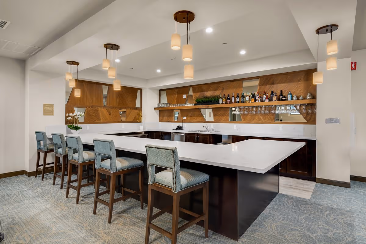 Modern communal kitchen/bar area with a large white island countertop, six upholstered bar stools, pendant lights, and shelving stocked with bottles and glassware.