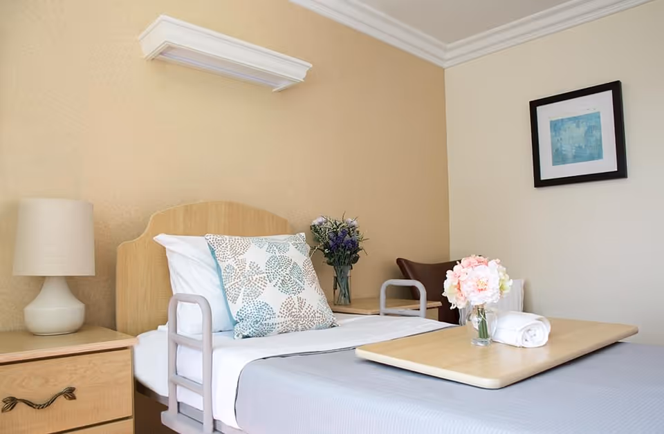 Neatly made single bed in a senior living room with bedside tables, a lamp, floral arrangements, and an overbed tray.