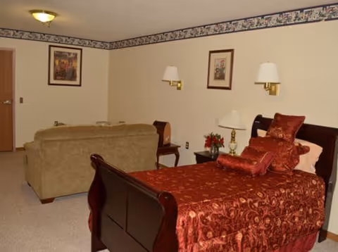 A furnished bedroom with a single wooden bed dressed in red bedding, a beige sofa, side tables, and wall lamps.