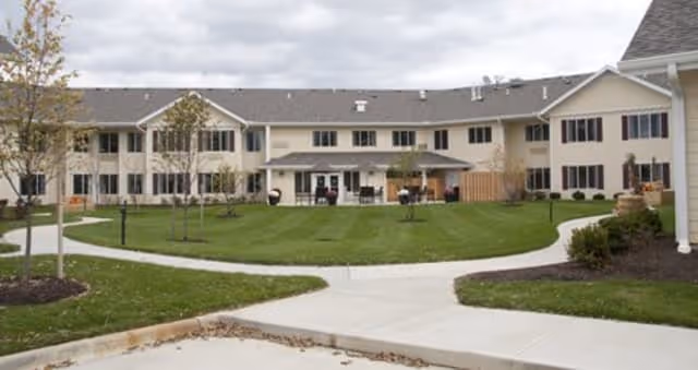Exterior view of a two-story senior living facility building with beige siding and multiple windows. There is a well-maintained lawn with young trees and a curved concrete walkway in front of the building under a cloudy sky.