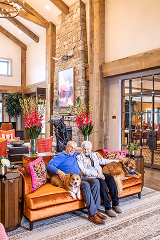 Two older adults seated on an orange sofa with two dogs in a bright, wood‑beamed common room featuring a stone fireplace and floral arrangements.