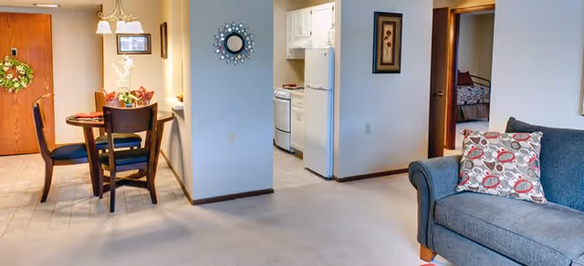 Interior view of a senior living facility showing a dining area with a round table and four chairs, a small kitchen with white appliances, and a living room with a gray couch and patterned pillow. The walls are light-colored, and there is a decorative mirror and framed artwork on the walls.