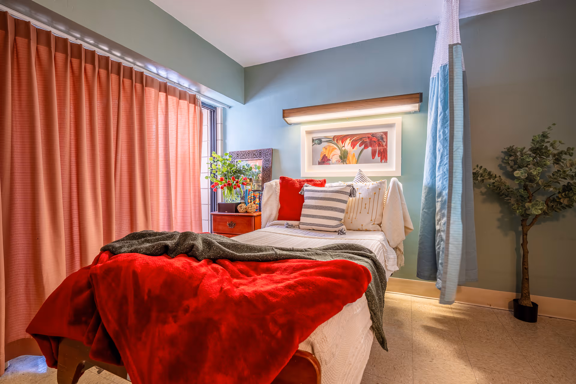 A cozy bedroom with a single bed covered in white bedding, a red blanket, and multiple pillows. There is a wooden nightstand with a vase of flowers and decorative items next to the bed. A colorful framed artwork is mounted on the wall above the bed, illuminated by a wall light. The room has green walls, a window with closed coral-colored curtains, a blue and white privacy curtain hanging near the bed, and a potted plant in the corner.