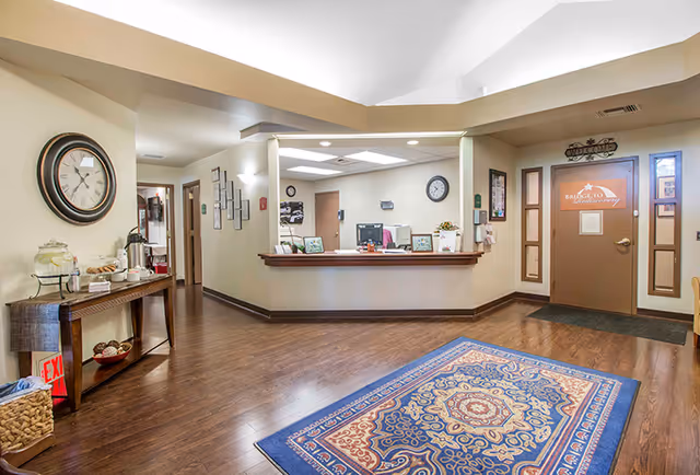 Reception area of a senior living facility with a wooden floor, a patterned blue and red rug, a wooden table with refreshments including a water dispenser and cups, a large wall clock, and a reception desk with computer monitors. There is a door with a sign that reads 'Bridge to Independence' and a 'Welcome' sign above it.