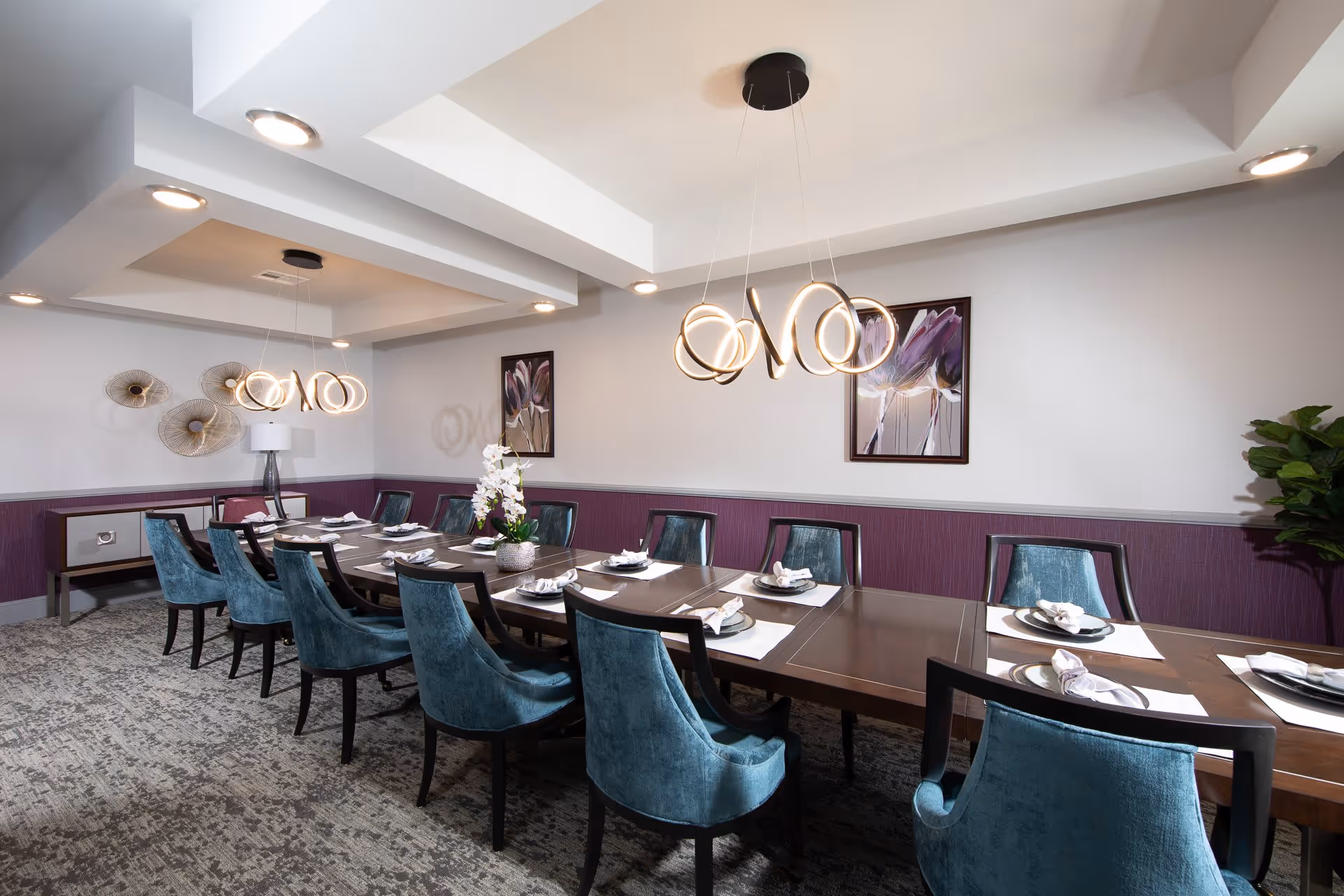 A modern dining room with a long wooden table set for a meal, surrounded by blue upholstered chairs. The room features contemporary circular pendant lights hanging from the ceiling, purple accent walls with floral artwork, a sideboard with decorative items, and a carpeted floor.