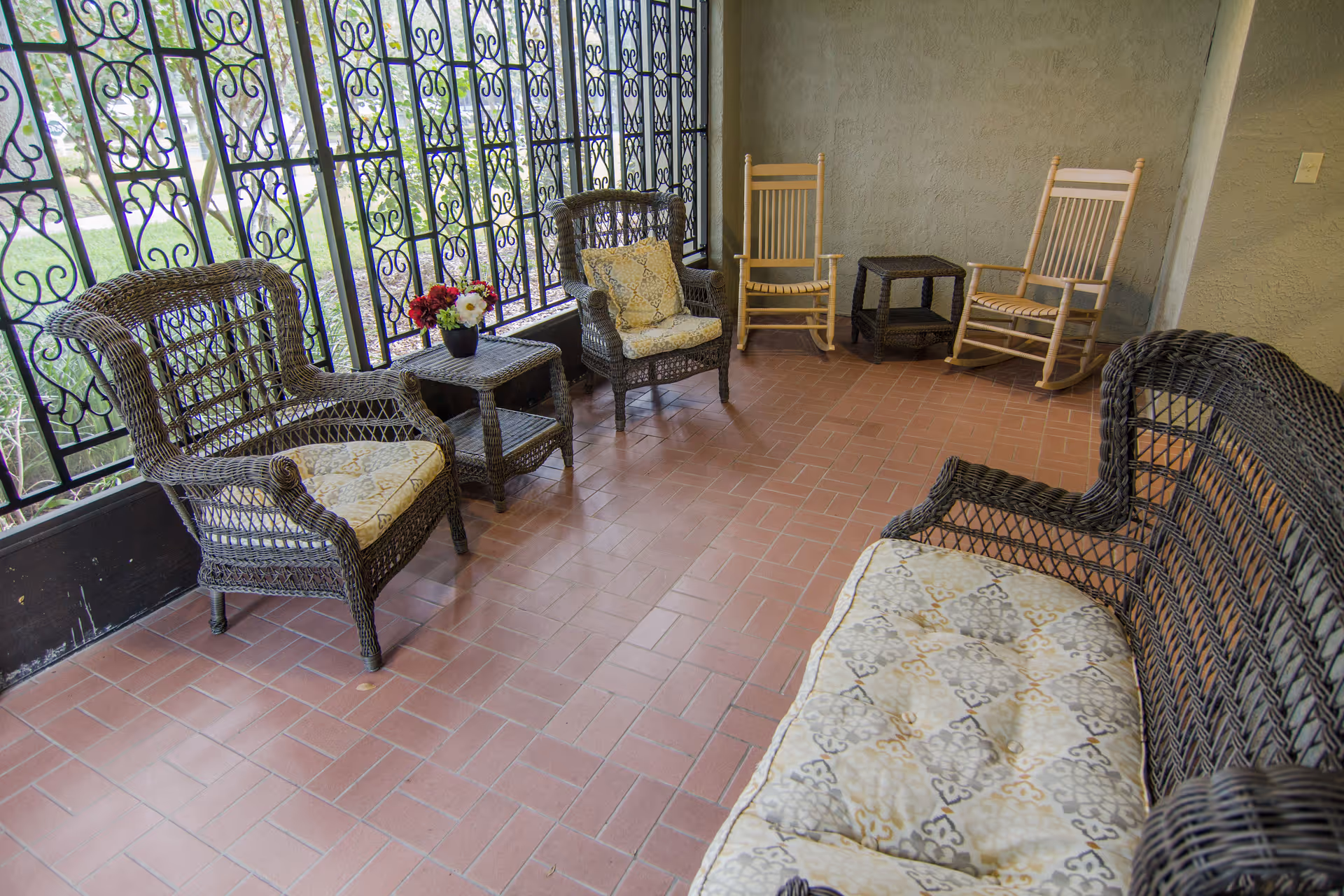 A cozy indoor seating area with wicker furniture including a cushioned sofa, two cushioned armchairs, two wooden rocking chairs, and two small wicker side tables. One side table holds a small pot of colorful flowers. The area has a tiled floor and decorative wrought iron window grilles with a view of greenery outside.