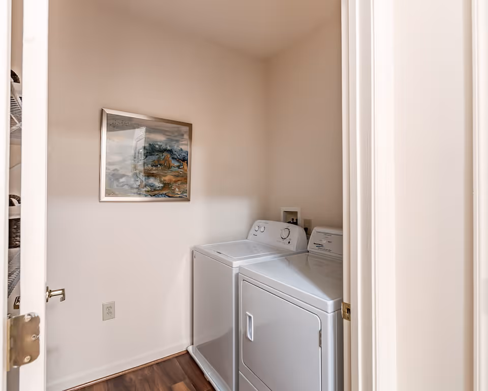 Small laundry room with a white washing machine and dryer side by side against a beige wall. A framed abstract painting hangs above the machines. The room has wooden flooring and is viewed through an open door.