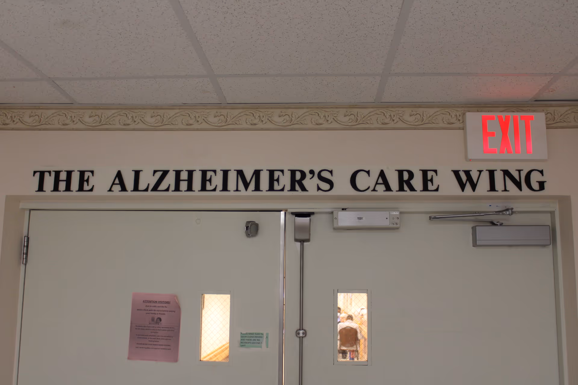 Double doors with small windows labeled 'THE ALZHEIMER'S CARE WING' above them. There is an illuminated red EXIT sign above the right door. Notices are posted on the left door.