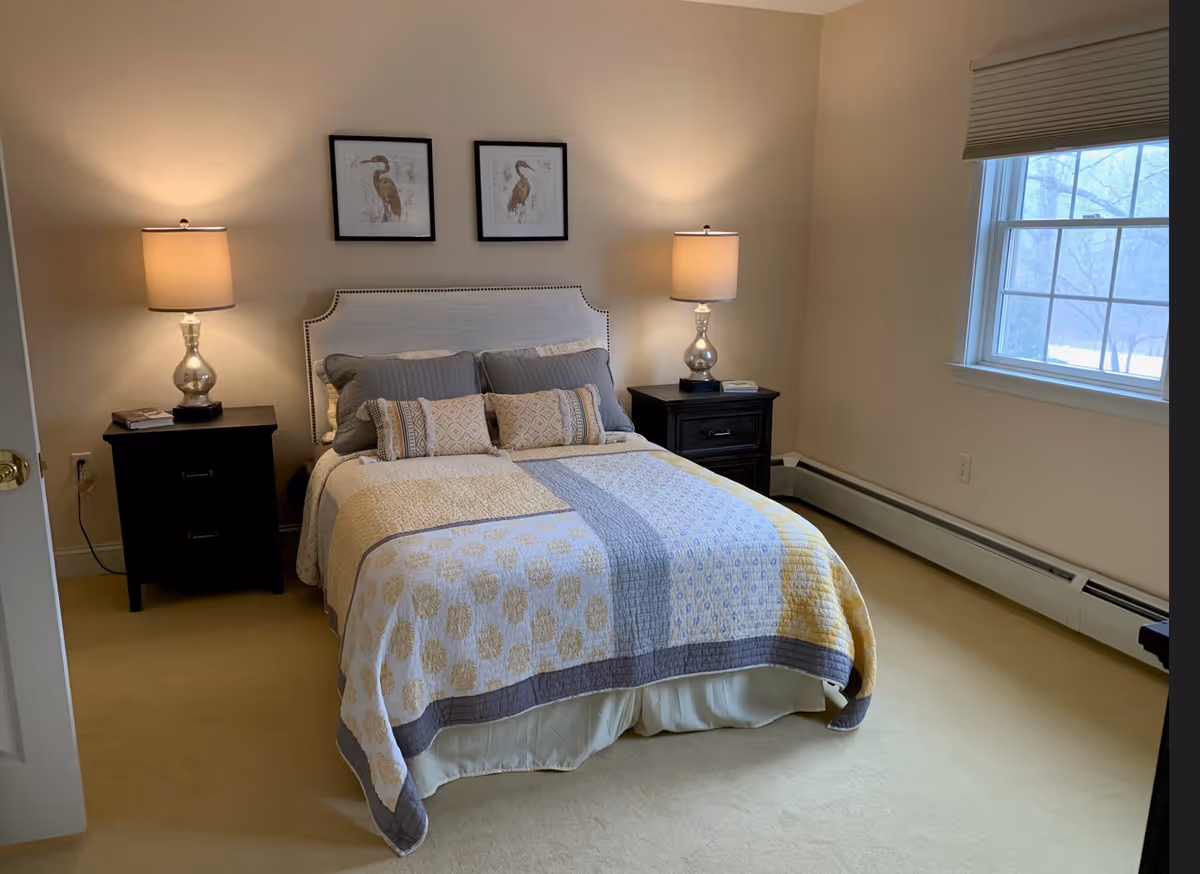 A neatly made bed with a patterned quilt and multiple pillows in a bedroom. There are two matching bedside tables with lamps on each side of the bed. Two framed bird pictures hang on the wall above the headboard. A window with blinds is on the right side of the room, letting in natural light.