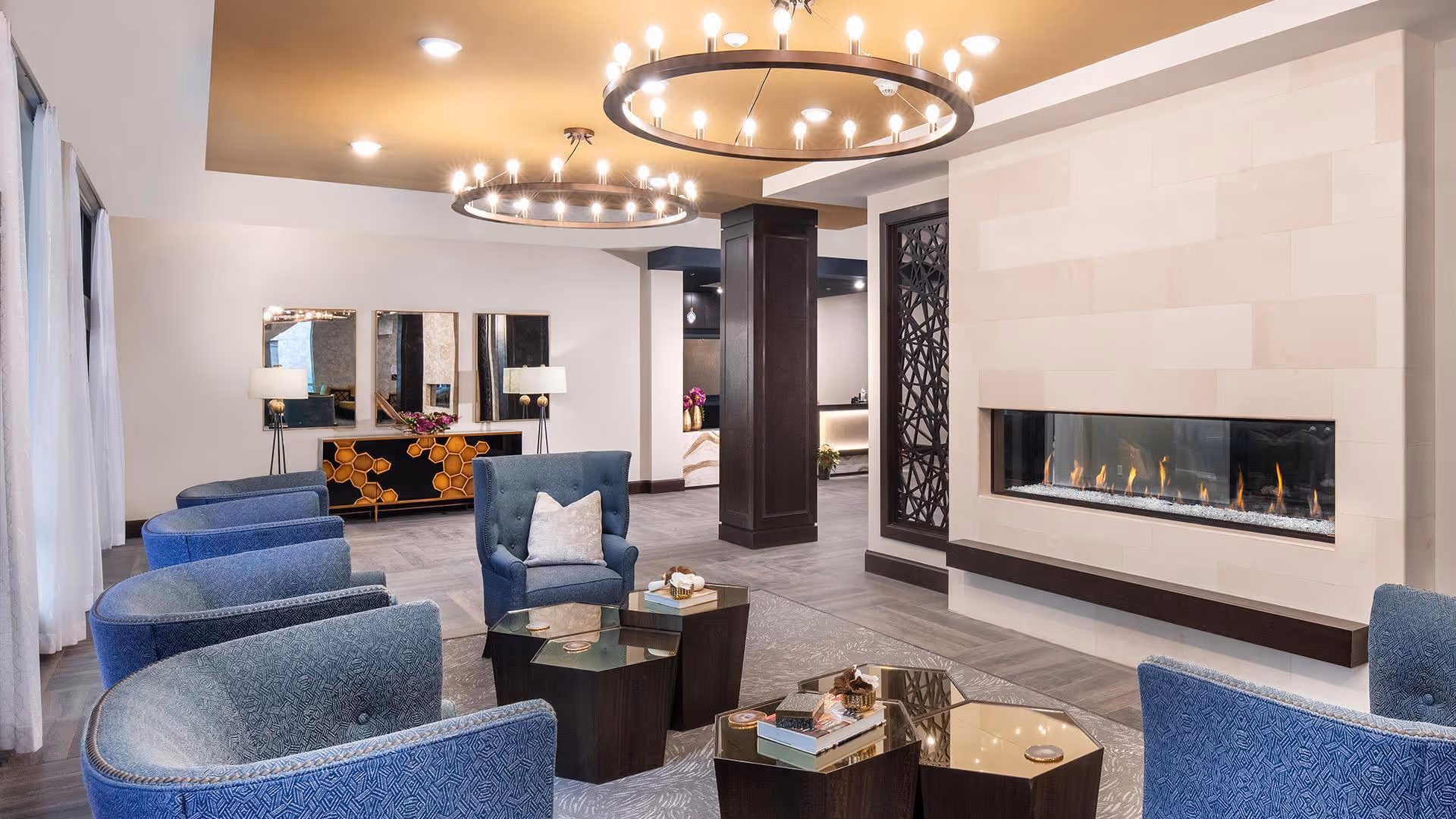 A modern and elegant living room area with blue upholstered armchairs arranged around a set of hexagonal glass-top coffee tables. A long, rectangular gas fireplace is built into a light-colored tiled wall. Two large circular chandeliers with multiple small lights hang from the ceiling. In the background, there is a console table with a honeycomb pattern and three mirrors above it, flanked by two lamps. The floor is covered with large gray tiles and a patterned area rug.