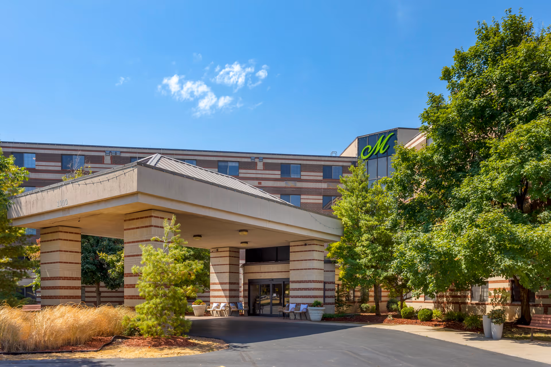 Front entrance of a multistory senior living building with a covered porte-cochere, landscaping, and a green "M" logo on the facade.