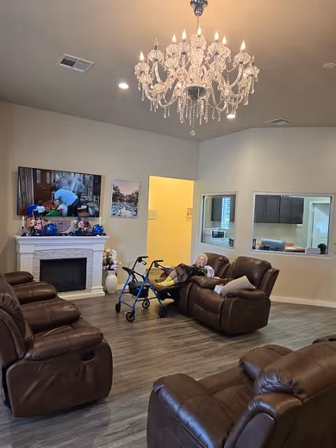 A cozy living room with multiple brown leather recliners arranged around a white fireplace. A flat-screen TV is mounted above the fireplace, showing a scene with a person. A chandelier hangs from the ceiling. A woman is sitting on one of the recliners with a blue walker in front of her. The room has wood flooring and light-colored walls with two windows looking into another room.