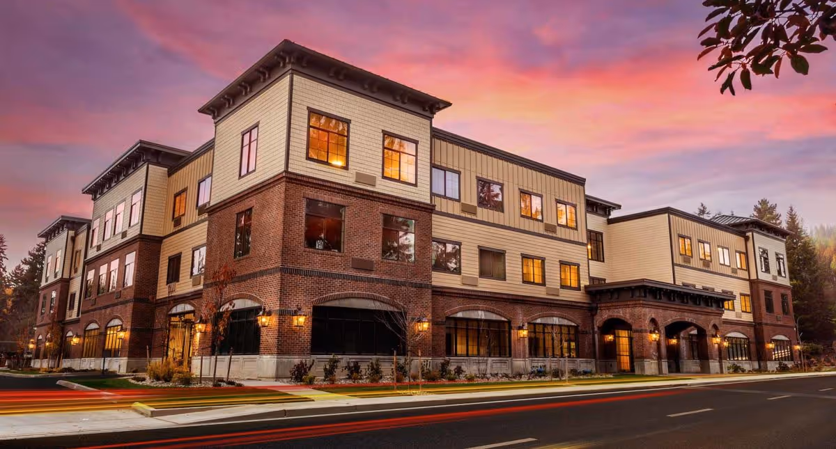 Exterior view of a three-story senior living facility building with brick and beige siding under a colorful sunset sky. The building has multiple windows with warm lights glowing inside and outdoor wall lanterns illuminating the entrance and surrounding area. Trees and landscaping are visible around the building.