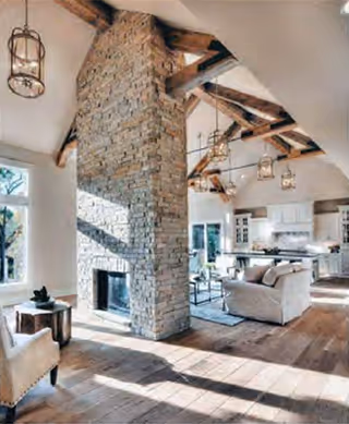 Bright open living area with a floor-to-ceiling stone fireplace, exposed wooden beams, seating, and a kitchen in the background.