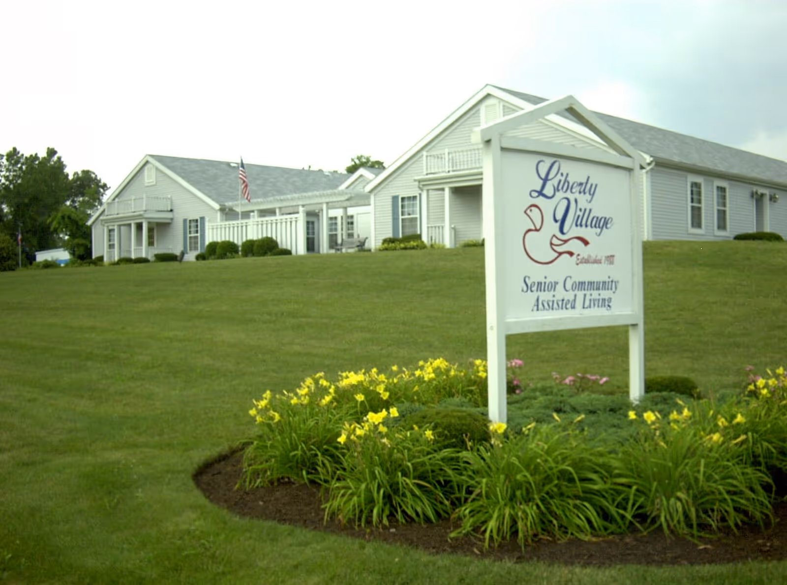 Exterior view of Liberty Village Senior Community building with a large green lawn and flower bed featuring yellow flowers in the foreground. A white sign with the community name and logo is prominently displayed near the flower bed.