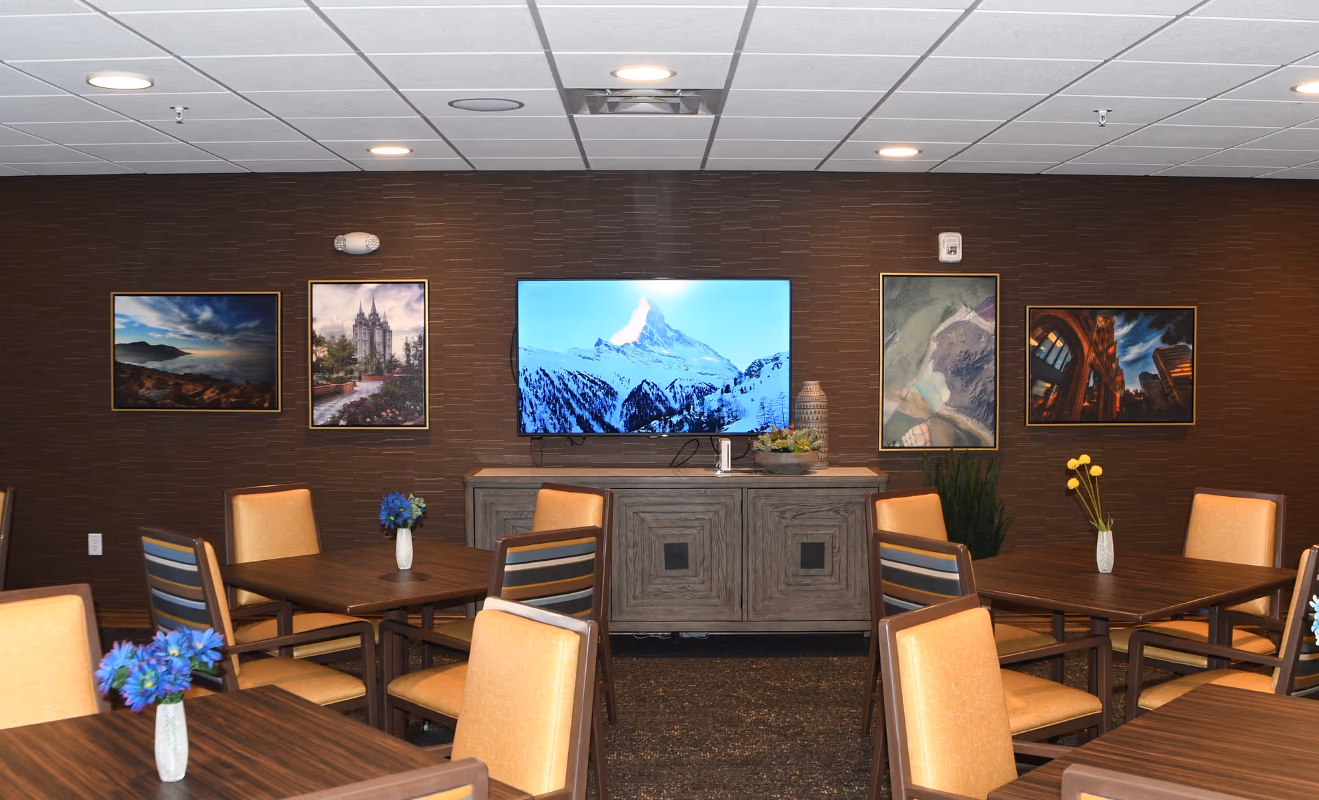 Interior view of a common area with several wooden tables and chairs with orange cushions. Each table has a small vase with flowers. A wooden cabinet against a brown textured wall holds a large flat-screen TV displaying a snowy mountain scene. The wall is decorated with five framed pictures of various landscapes and buildings.