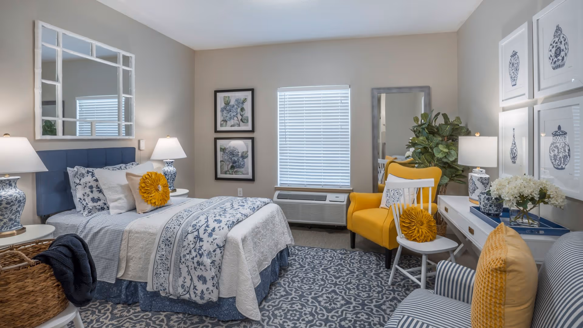 A cozy bedroom with a blue and white color scheme featuring a bed with floral bedding and a yellow decorative pillow. The room has two bedside tables with matching blue and white lamps, a large mirror above the bed, framed botanical prints on the wall, a yellow armchair, a white chair with a yellow pillow, a striped armchair, a white desk with a blue tray and flowers, and a patterned blue and white rug on the floor.