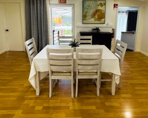 A dining area with a rectangular table covered with a white tablecloth and six wooden chairs around it. There is a small plant centerpiece on the table. The room has wooden flooring, a white brick fireplace with a decorative painting above it, and an exit door with a window showing an outdoor area. Curtains are partially drawn on the left side of the door.