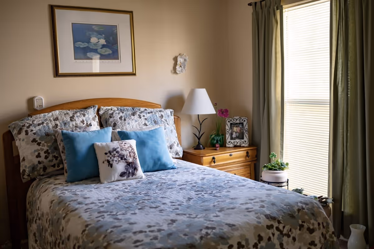 A cozy bedroom with a bed covered in a floral-patterned quilt and several pillows, including two blue ones and one with an owl design. Next to the bed is a wooden nightstand with a white lampshade, a framed photo, and a small potted plant. A window with green curtains and blinds allows natural light into the room. A framed picture hangs on the beige wall above the bed.