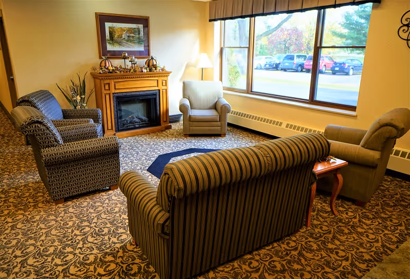 A cozy sitting room with a sofa and armchairs arranged around a fireplace beside a large window overlooking a parking lot.