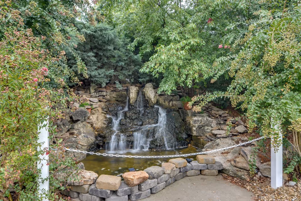 A serene outdoor garden area featuring a small cascading waterfall flowing over rocks into a pond, surrounded by lush green trees and bushes. A low stone wall and a white chain barrier frame the pond area.