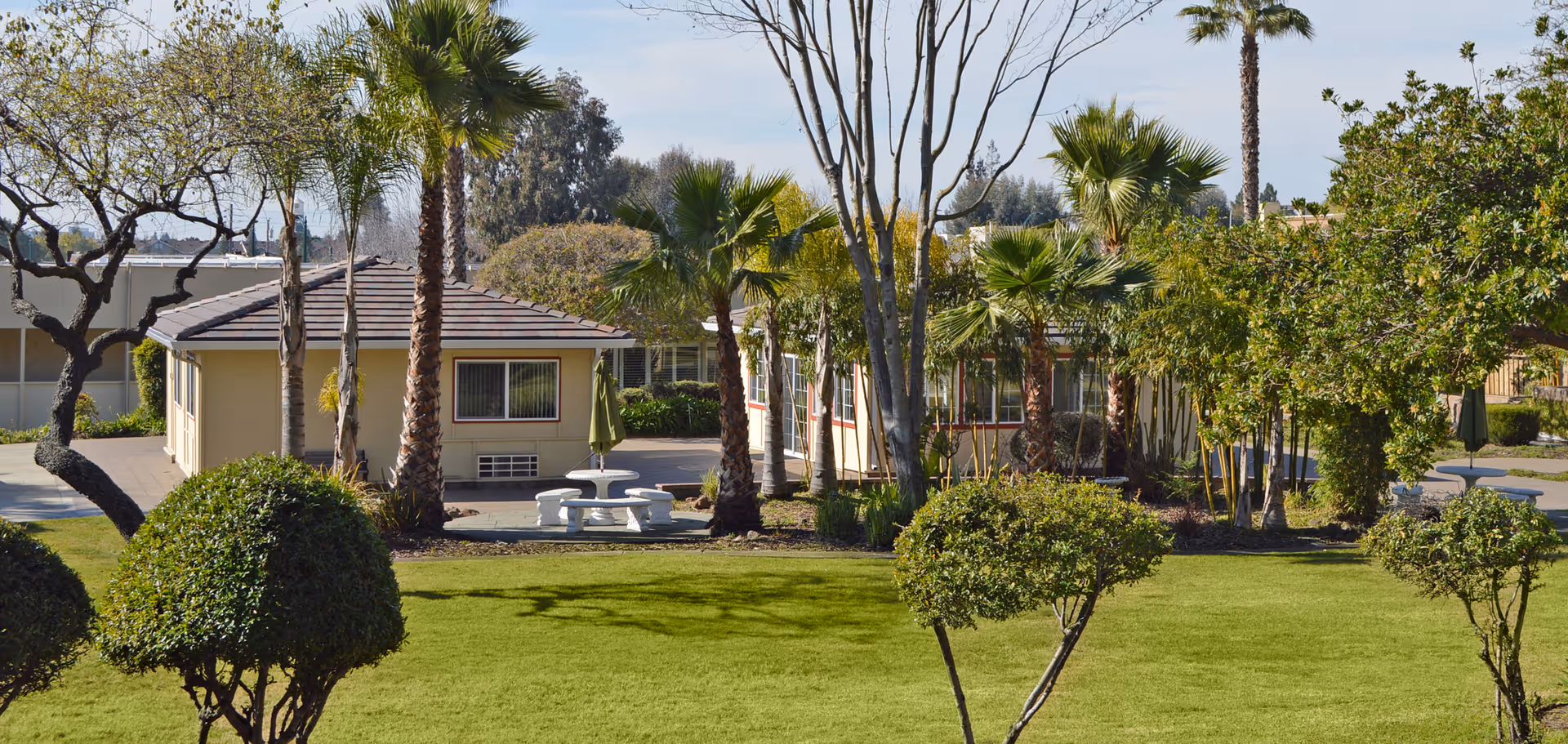 Landscaped courtyard with a green lawn, palm trees, small single-story buildings, and a white outdoor picnic table.