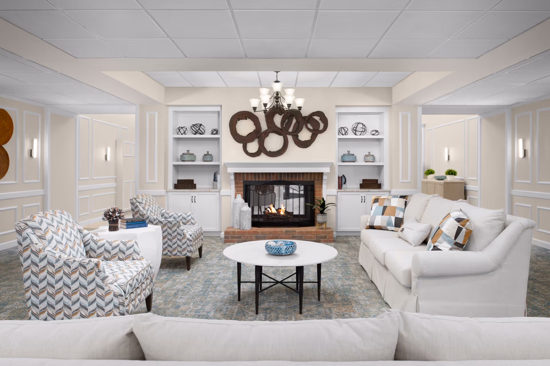 A cozy living room with a lit brick fireplace in the center, flanked by built-in white shelves decorated with vases and ornamental spheres. In front of the fireplace is a round white coffee table with a blue decorative bowl. The seating includes a white sofa with patterned pillows on the right and two patterned armchairs on the left. The room has beige walls with white trim and soft lighting from wall sconces and a chandelier above the fireplace.