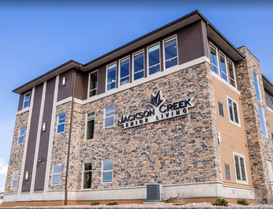 Exterior view of a multi-story building with stone and brown siding, featuring large windows and a sign that reads 'Jackson Creek Senior Living' on the front wall under a clear blue sky.