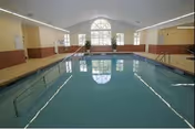 Indoor swimming pool with clear blue water in a well-lit room featuring a large arched window at the far end, beige walls with brown lower panels, handrails on both sides of the pool, and a tiled floor surrounding the pool area.