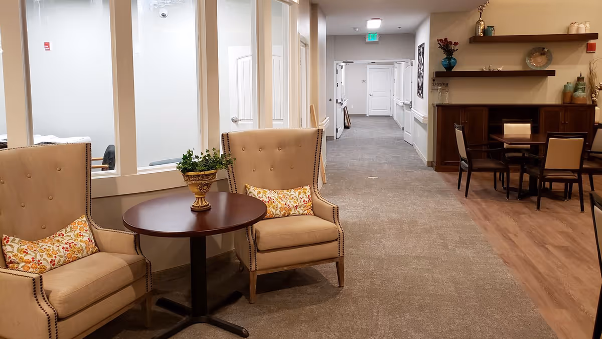 A cozy seating area in an assisted living facility hallway featuring two beige armchairs with floral cushions around a small round wooden table with a decorative plant. The hallway extends into the distance with carpeted flooring, and to the right is a dining area with a wooden floor, table, and chairs, along with shelves holding decorative items.