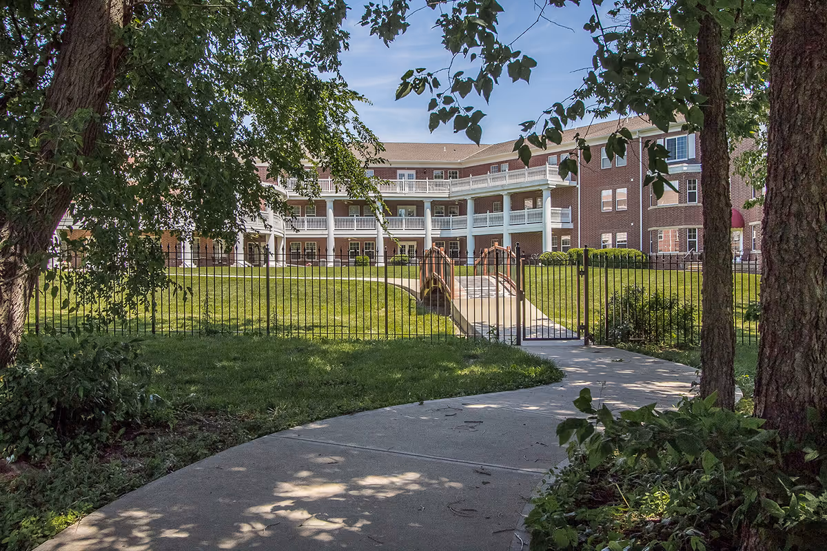 View of a senior living community building with a three-story brick facade and white balconies. In the foreground, there is a curved concrete pathway leading to a gated entrance with a small bridge over a grassy area. Trees and shrubs frame the pathway, providing shade and greenery.