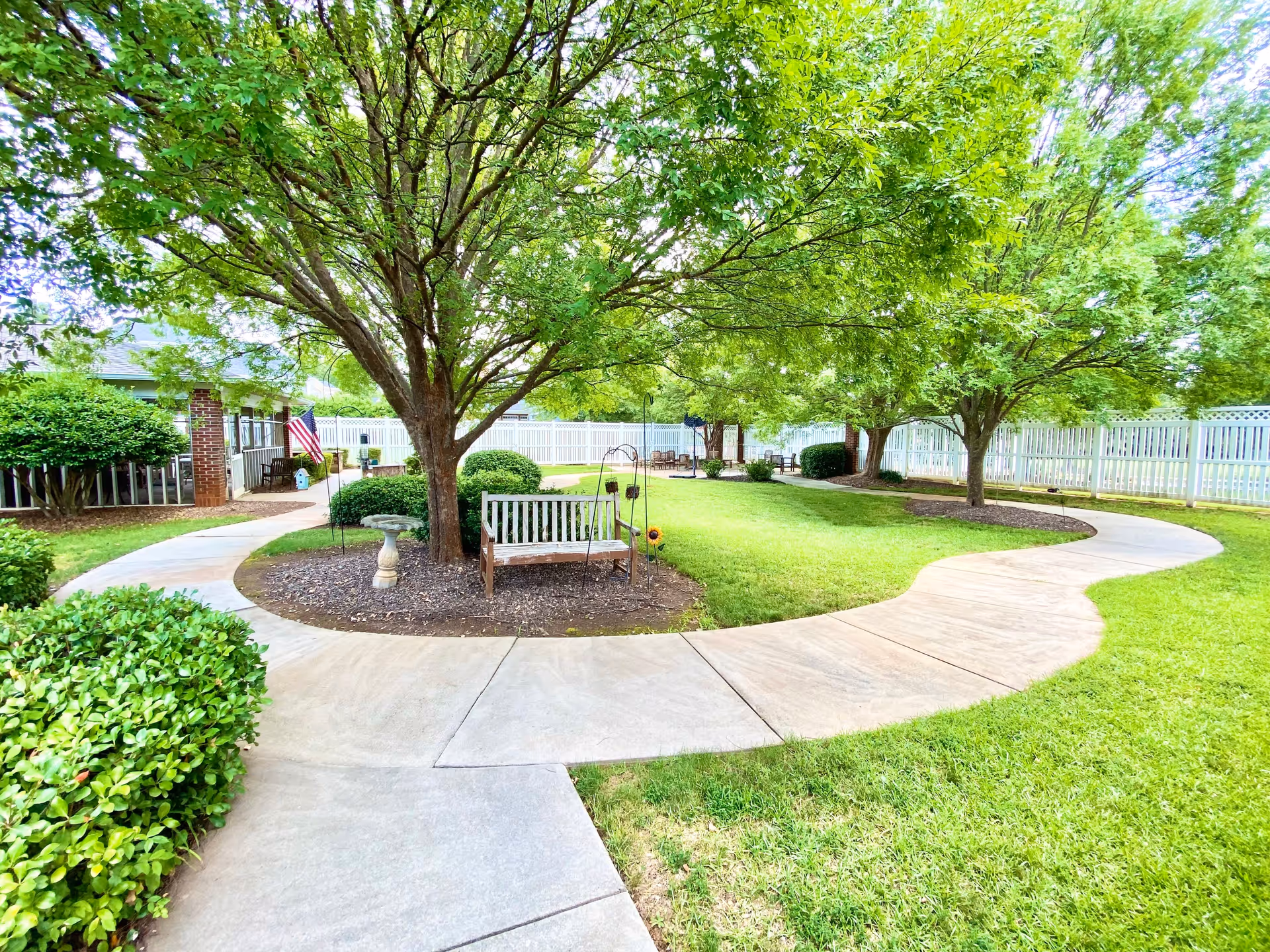 A well-maintained outdoor garden area with a curved concrete walkway, green grass, several trees providing shade, a white wooden bench, and a white fence enclosing the space. There is an American flag near a building on the left side.