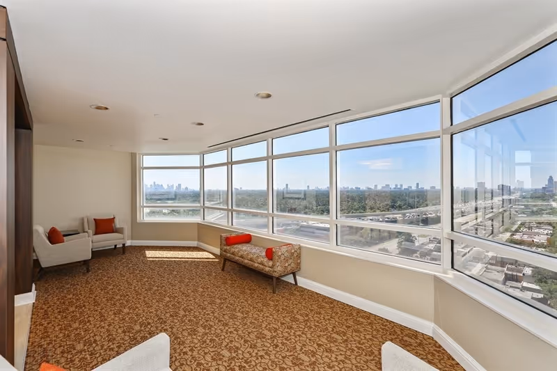 A bright, spacious lounge area with large floor-to-ceiling windows offering a panoramic view of a city skyline and greenery. The room features patterned carpet flooring, a floral upholstered bench with red cylindrical pillows, and several beige armchairs with red accent pillows.