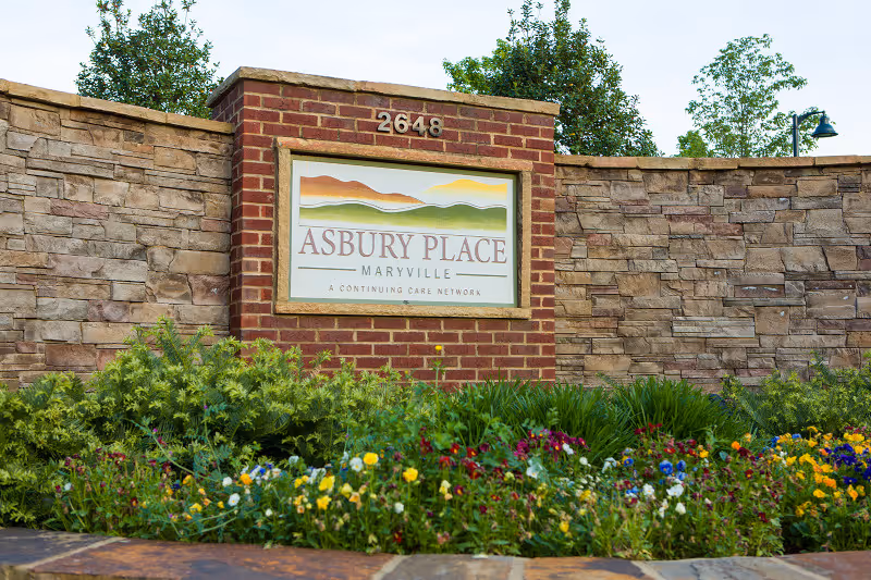 A brick and stone sign for Asbury Place Maryville, surrounded by green shrubs and colorful flowers, with trees and a lamp post in the background.