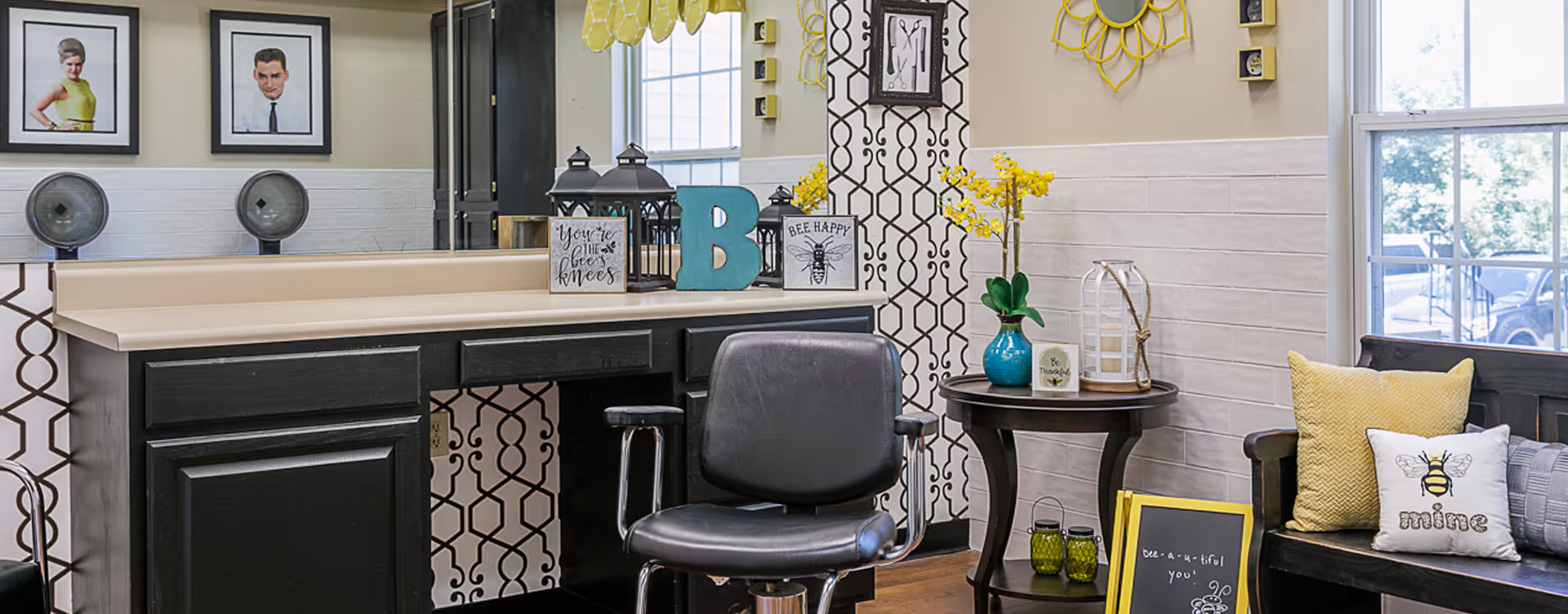 Interior of a salon area with a black salon chair in front of a black vanity with a large mirror. The vanity countertop has decorative items including lanterns, a large blue letter B, and signs with bee-themed sayings. The wall behind the vanity has patterned wallpaper and framed pictures. To the right, there is a small round table with a blue vase holding yellow flowers, a white lantern, and a small sign. A bench with yellow and bee-themed pillows is next to a window showing a view outside.