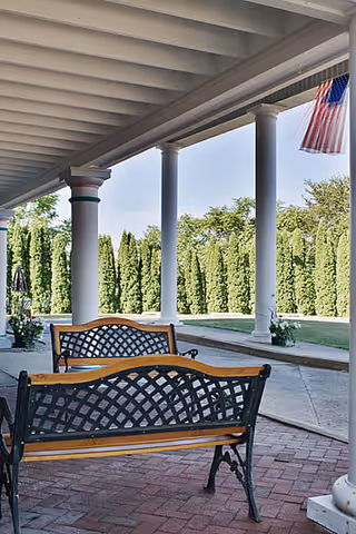 Covered outdoor patio area with two black metal and wood benches facing away, white columns supporting the roof, an American flag hanging, and tall green hedges in the background.
