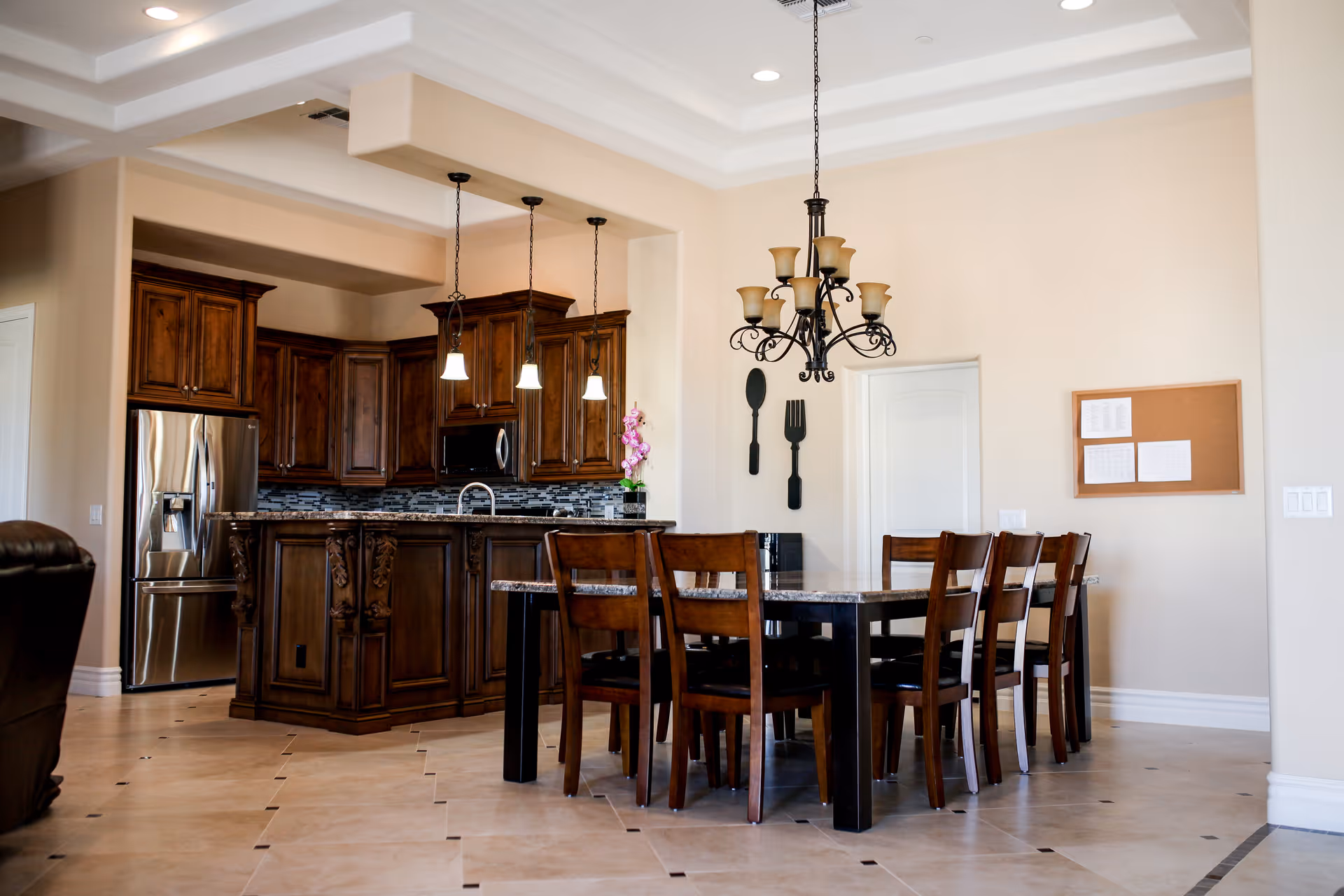 A spacious dining area with a dark wooden dining table and six matching chairs. Above the table hangs an ornate chandelier with multiple lights. In the background, there is a kitchen with dark wooden cabinets, a stainless steel refrigerator, a microwave, and a kitchen island with three pendant lights. The walls are painted beige, and there are decorative oversized fork and spoon wall hangings near a white door. The floor is tiled with a light beige color and dark accents.