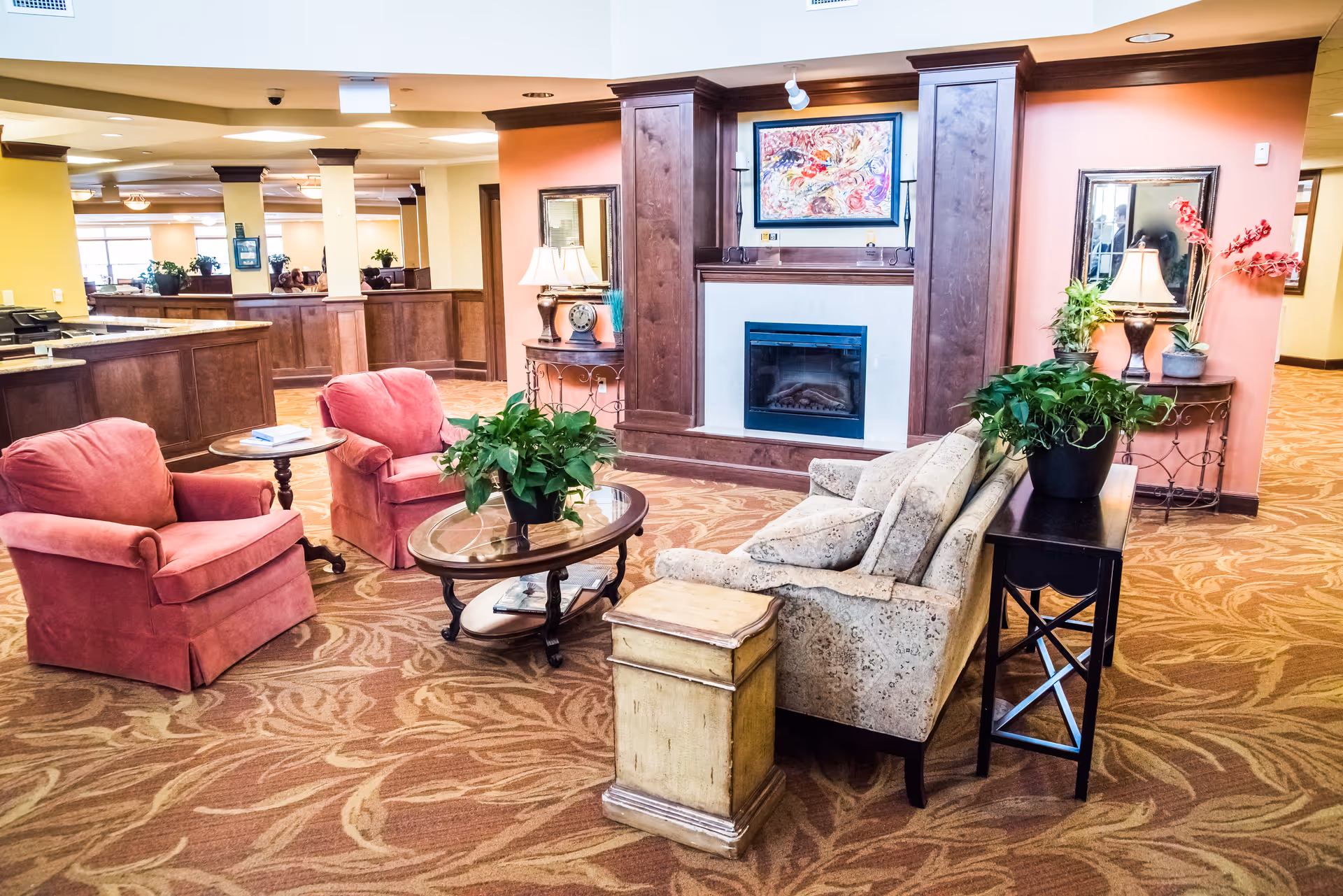 A cozy senior living facility common area with a fireplace surrounded by wooden paneling. The room features two red armchairs, a patterned beige sofa, a glass-top coffee table with a green plant, and side tables with lamps and decorative items. The carpet has a leaf pattern, and the walls are painted in warm tones with framed artwork and mirrors.